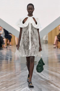Runway model walking a catwalk in a white lace knee-length dress with a dramatic white cape collar and bow, carrying a green quilted handbag.