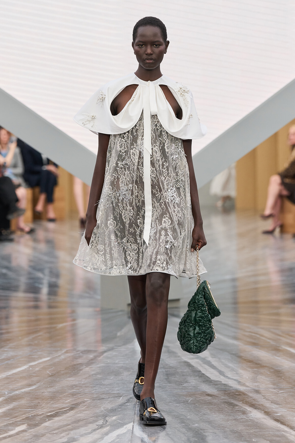 Runway model walking a catwalk in a white lace knee-length dress with a dramatic white cape collar and bow, carrying a green quilted handbag.