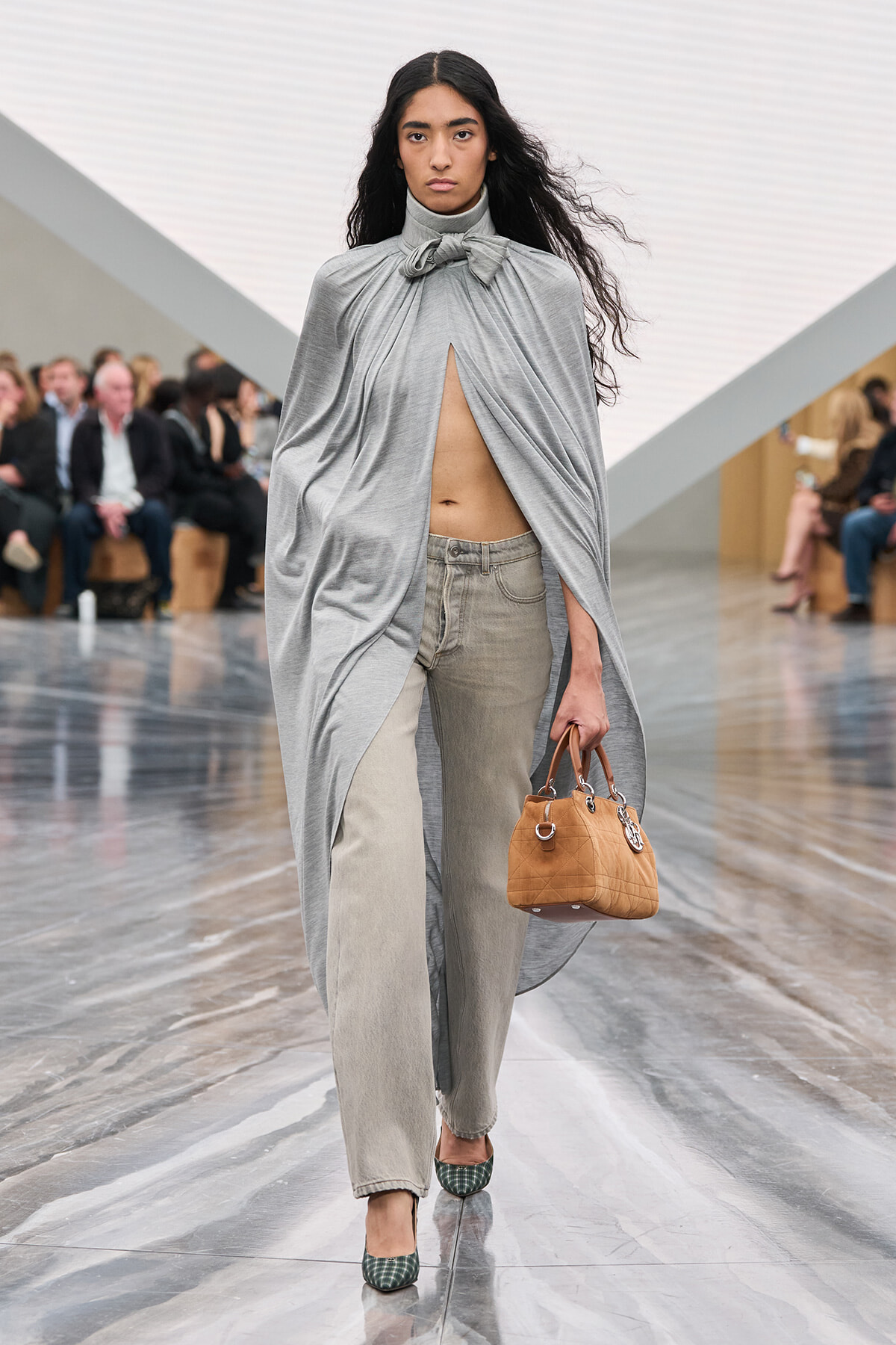 Model walking a runway in a light-gray draped cape tied at the neck, exposing midriff, paired with gray jeans and green plaid heels, carrying a tan quilted handbag.