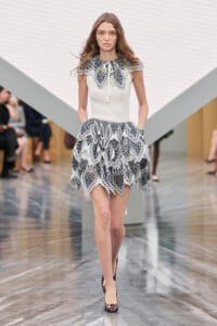 Model walking a runway in a cream knit top with a lace collar and a tiered black-and-white lace skirt, burgundy heels.