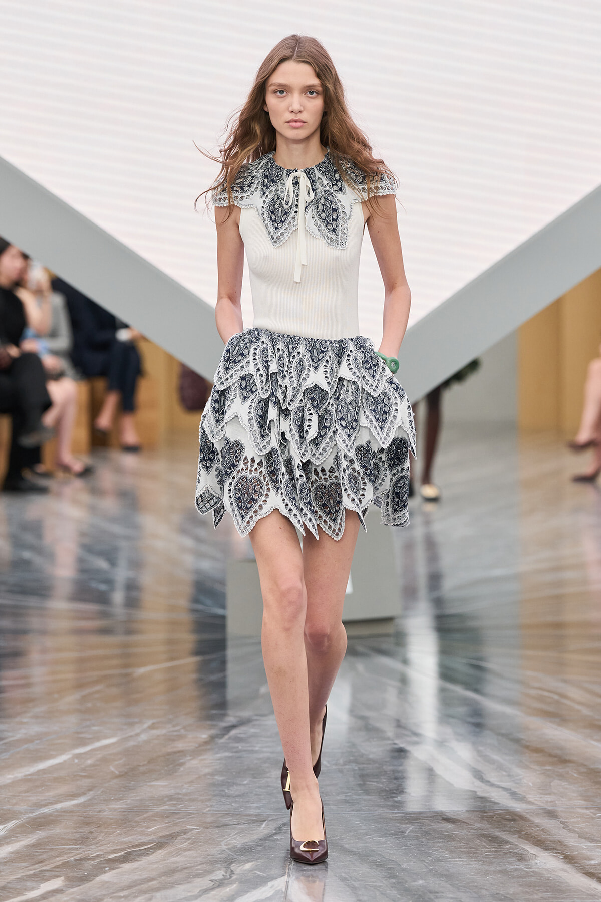 Model walking a runway in a cream knit top with a lace collar and a tiered black-and-white lace skirt, burgundy heels.