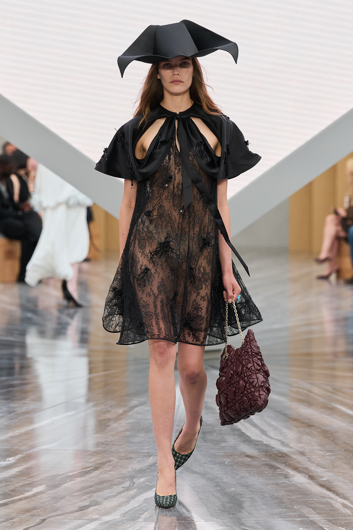 Model walking the runway in a sheer black lace dress with a dramatic black capelet and a large sculptural hat; holding a burgundy quilted bag with a chain strap
