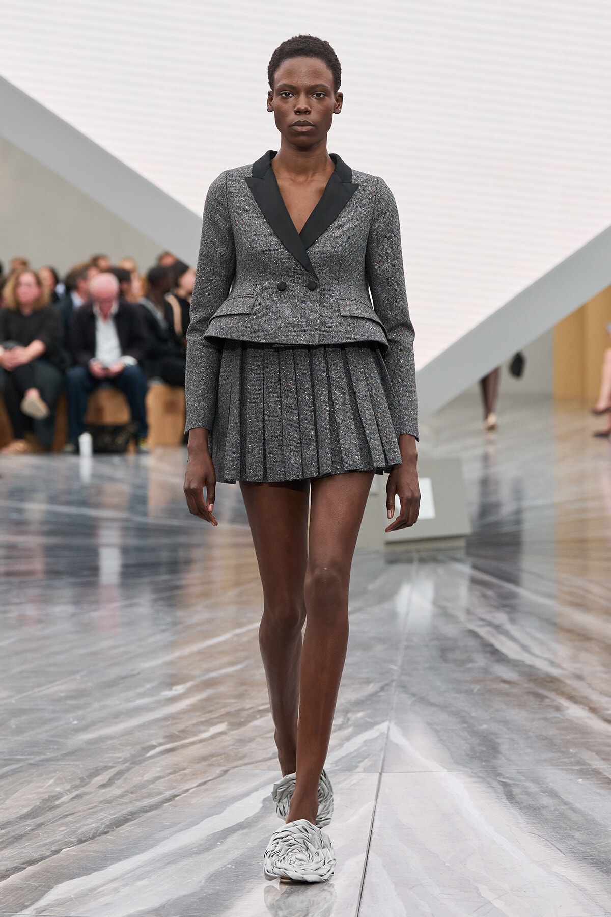 Model walking a fashion runway in a metallic gray double-breasted blazer with black lapels and a matching pleated mini-skirt, paired with white braided footwear; audience blurred in the background.