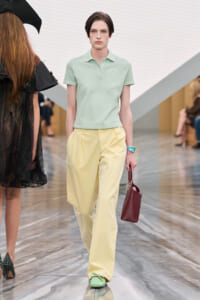 Model on a runway wearing a pastel mint polo, pale yellow trousers, mint shoes, turquoise bracelet, and a burgundy handbag.