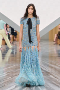 Model walks the runway in a light-blue beaded gown with short sleeves and a black neck bow.