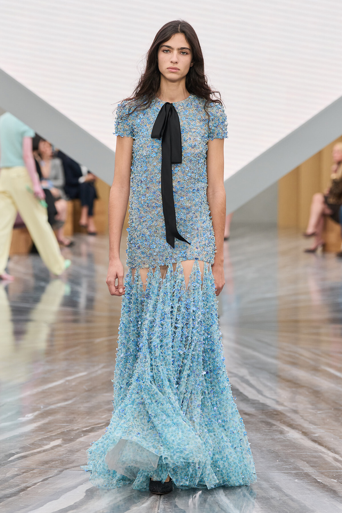 Model walks the runway in a light-blue beaded gown with short sleeves and a black neck bow.