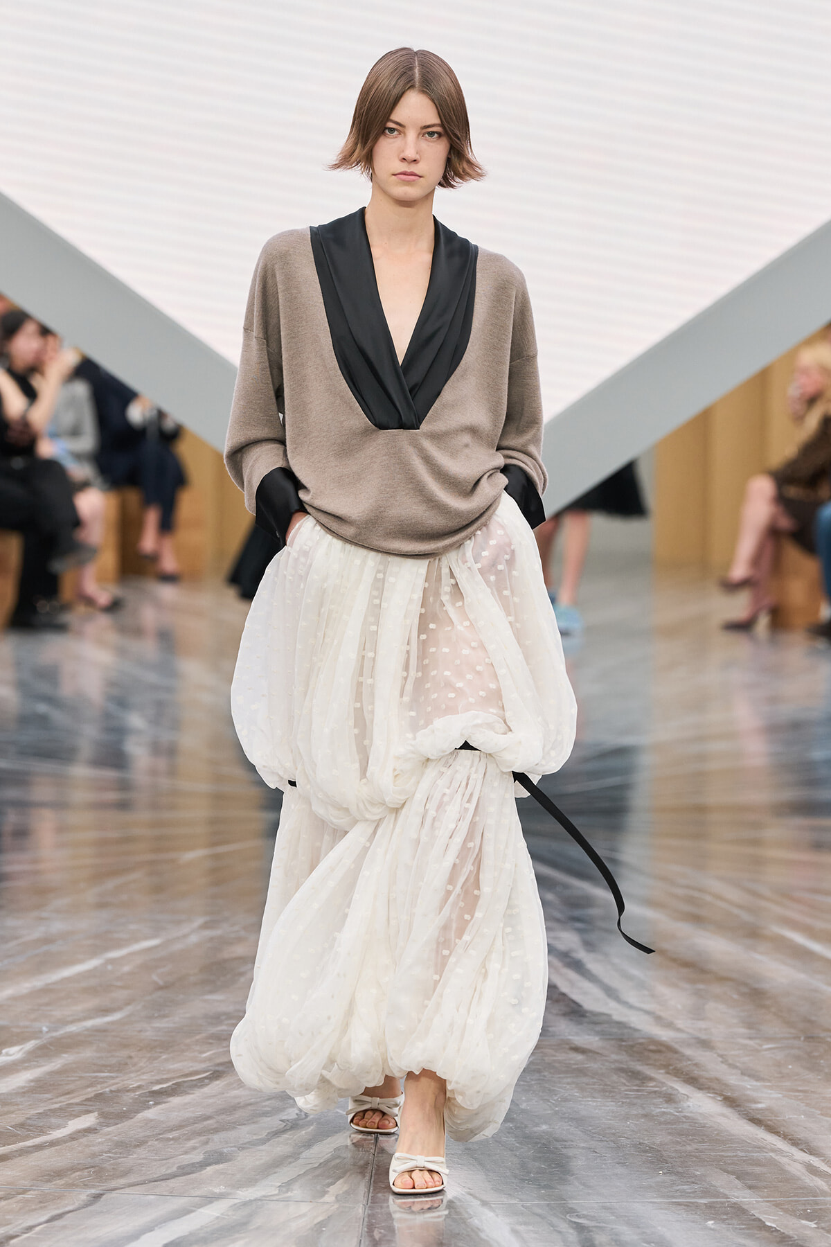 Model walking a runway in a taupe draped sweater with a black V-neck and a voluminous sheer white polka-dot skirt with pockets at the hips.