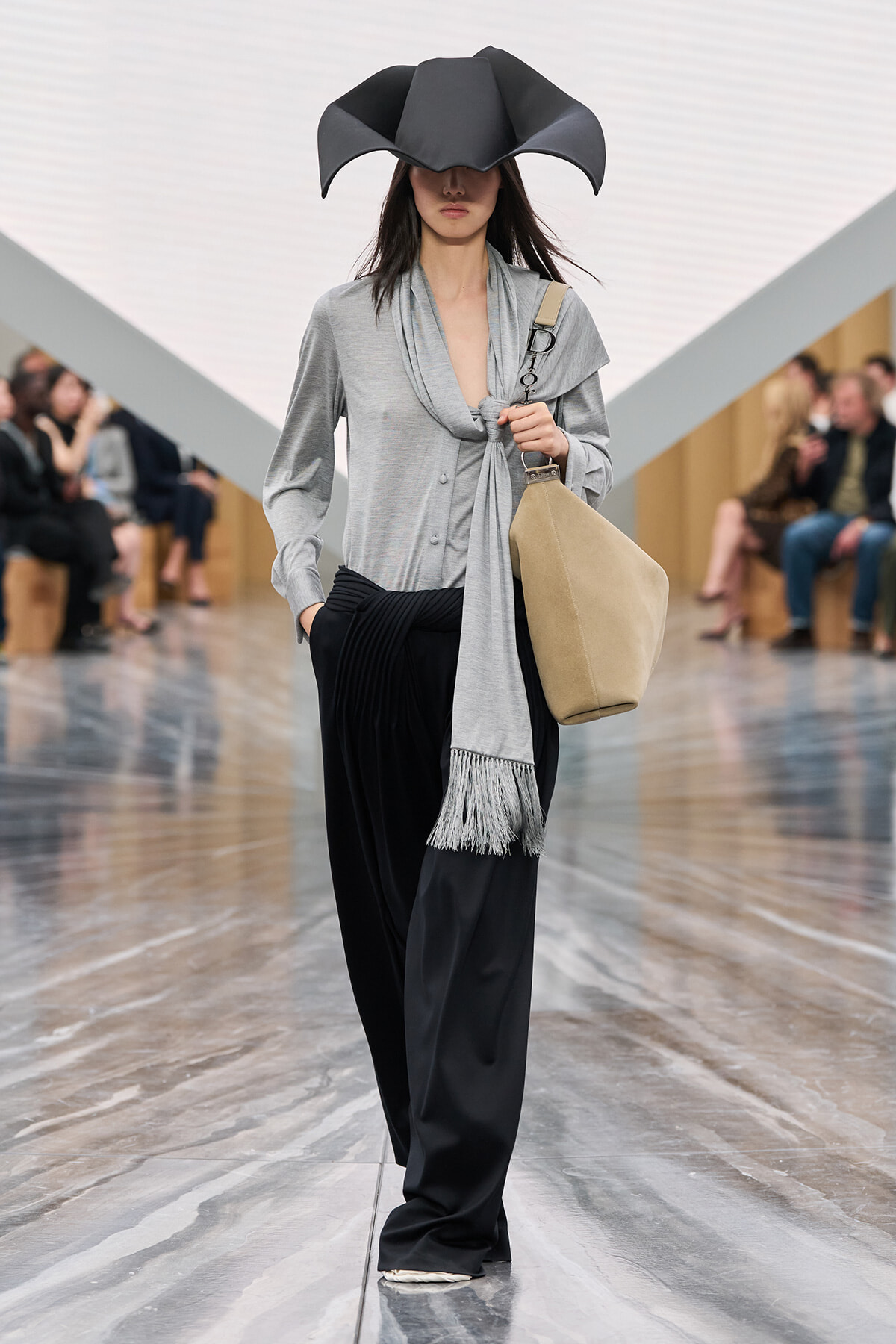Model walking a runway in an oversized black sculptural hat, gray wrap blouse with scarf, and black wide-leg pants with a beige handbag.