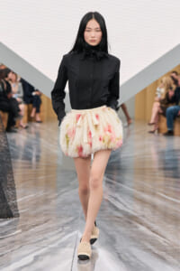 Model walking a runway in a black buttoned blouse with bow and a fluffy pastel skirt tied with a cream ribbon, beige slippers visible.
