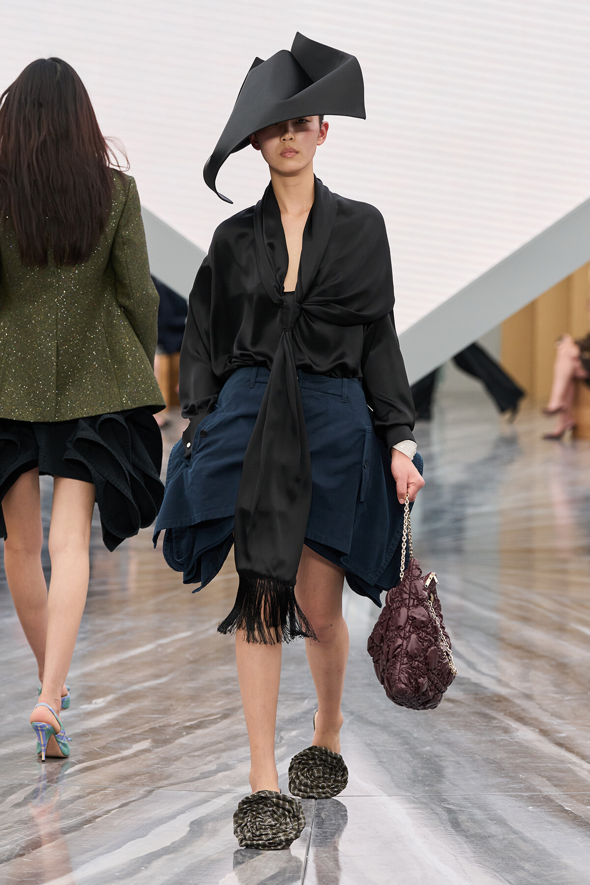 Runway model in a black satin blouse tied at the waist, navy layered skirt with fringe, an oversized sculptural black hat, and a quilted purple handbag with a chain strap.