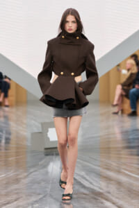 Runway model wearing a dark brown structured coat with cutouts at the waist, gold buttons, paired with a gray mini-skirt and green heeled sandals.