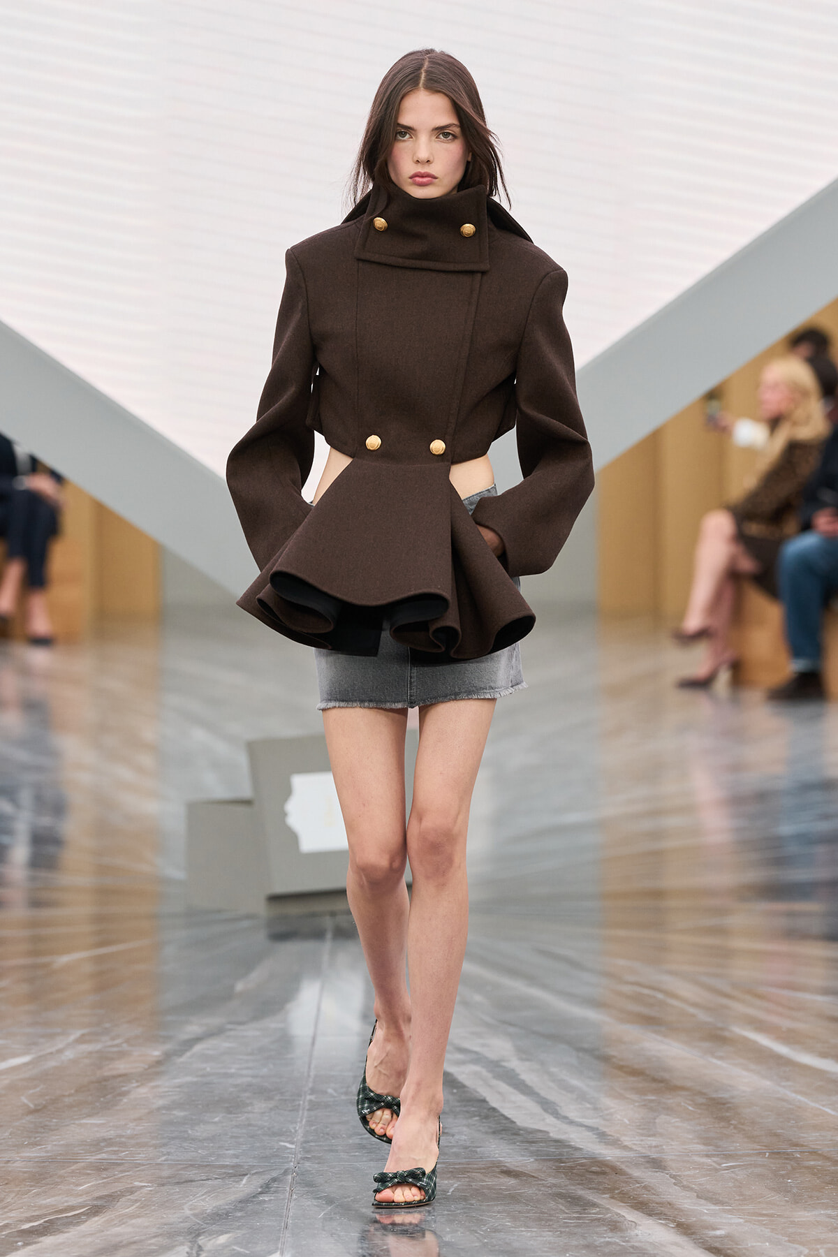 Runway model wearing a dark brown structured coat with cutouts at the waist, gold buttons, paired with a gray mini-skirt and green heeled sandals.