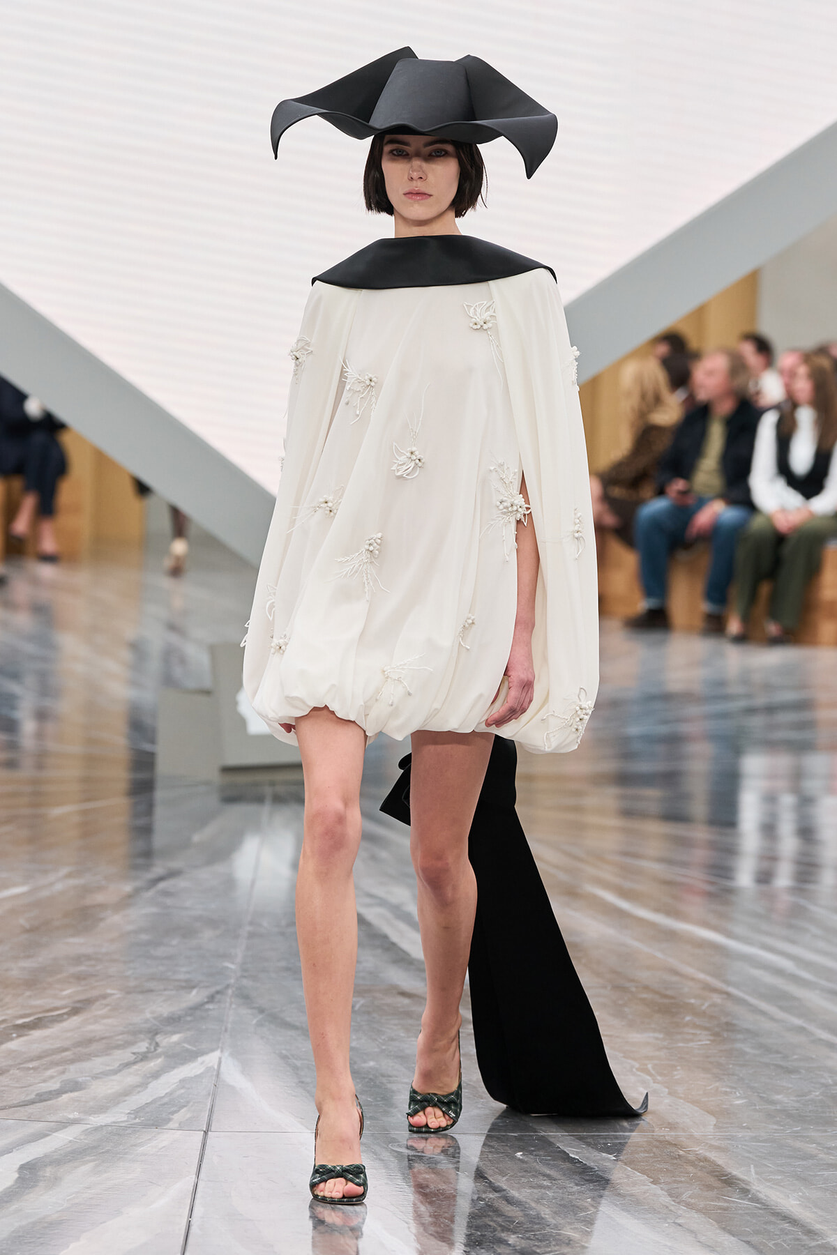 Model walks the runway in an ivory, voluminous cape dress with floral embroidery, paired with a sculptural black hat and trailing black cape behind her.