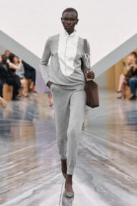 Model on a runway wearing a light gray knit suit with a white scalloped-front blouse and a brown shoulder bag.