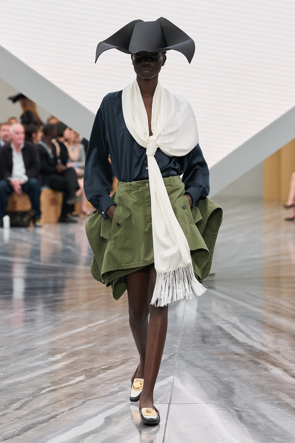 Model on a runway wearing a navy blouse, tied white scarf, and olive green oversized cargo-style skirt with big pockets, plus a dramatic black sculptural hat.