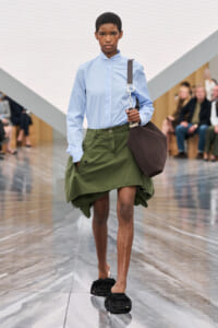 Model walking a fashion runway in a light blue high-collar shirt, olive oversized skirt, black rosette slippers, and a brown tote over the shoulder. Audience blurred in background.