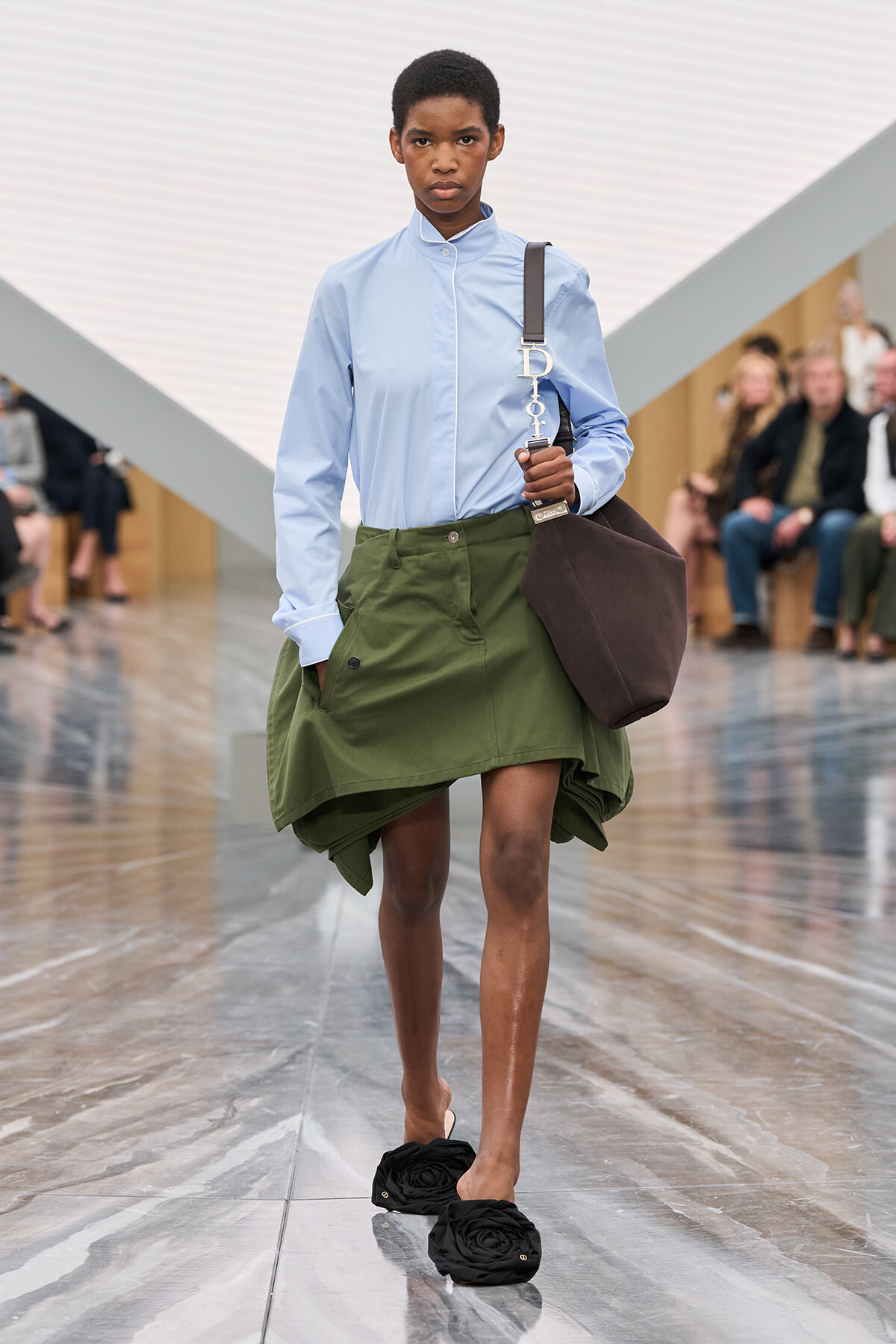 Model walking a fashion runway in a light blue high-collar shirt, olive oversized skirt, black rosette slippers, and a brown tote over the shoulder. Audience blurred in background.