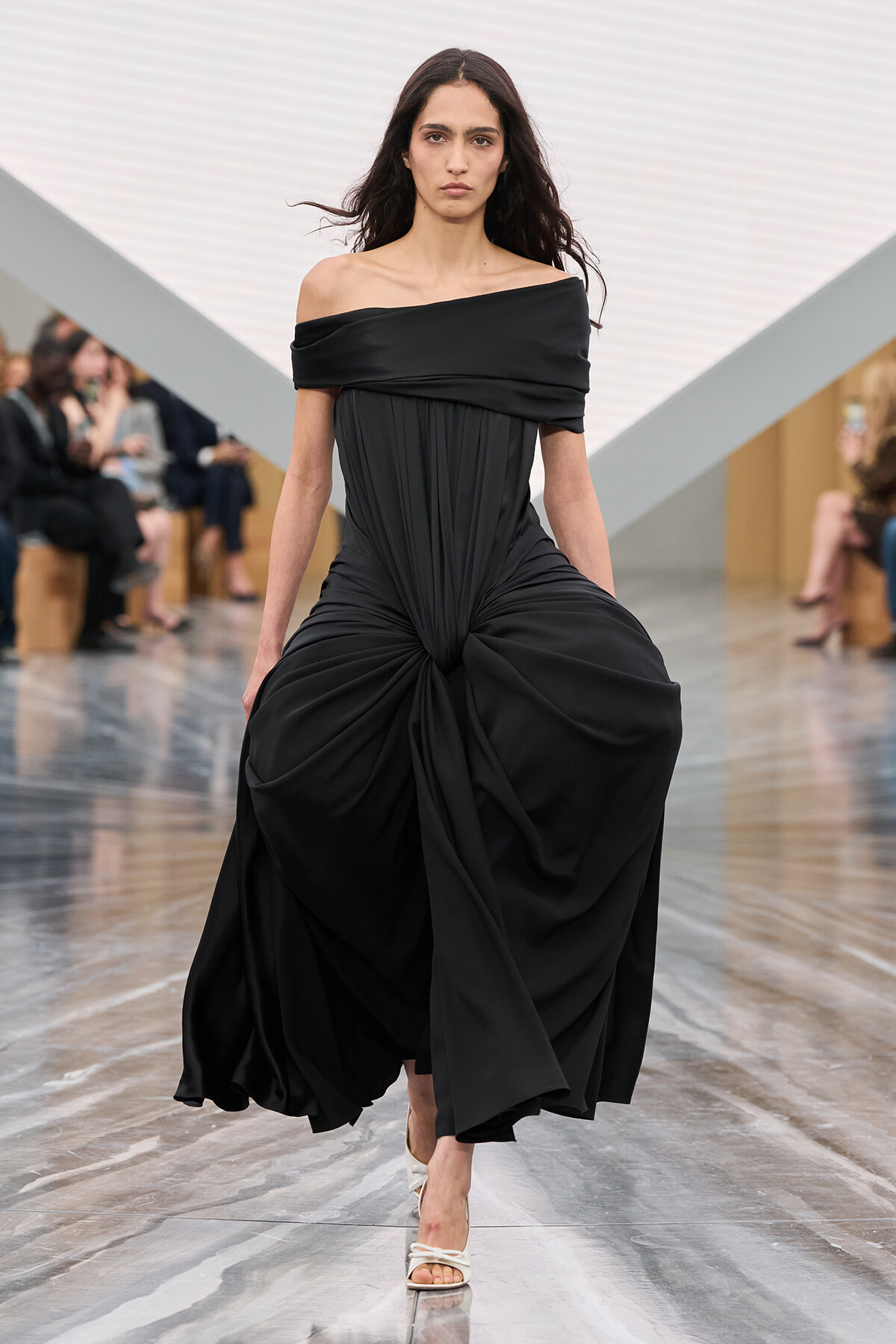 Model walking a runway in a black off-shoulder draped gown with a twisted knot at the waist, white heels, and a neutral expression.