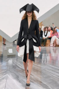 Runway model in a black tailored blazer with a deep V, oversized bow at the waist, and a white layered skirt with a large black sculptural hat above. Audience in background.