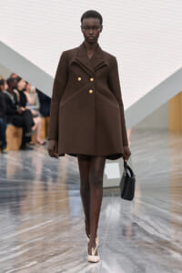 Model walks the runway in a brown structured double-breasted coat with gold buttons, carrying a small black handbag and wearing beige heels.