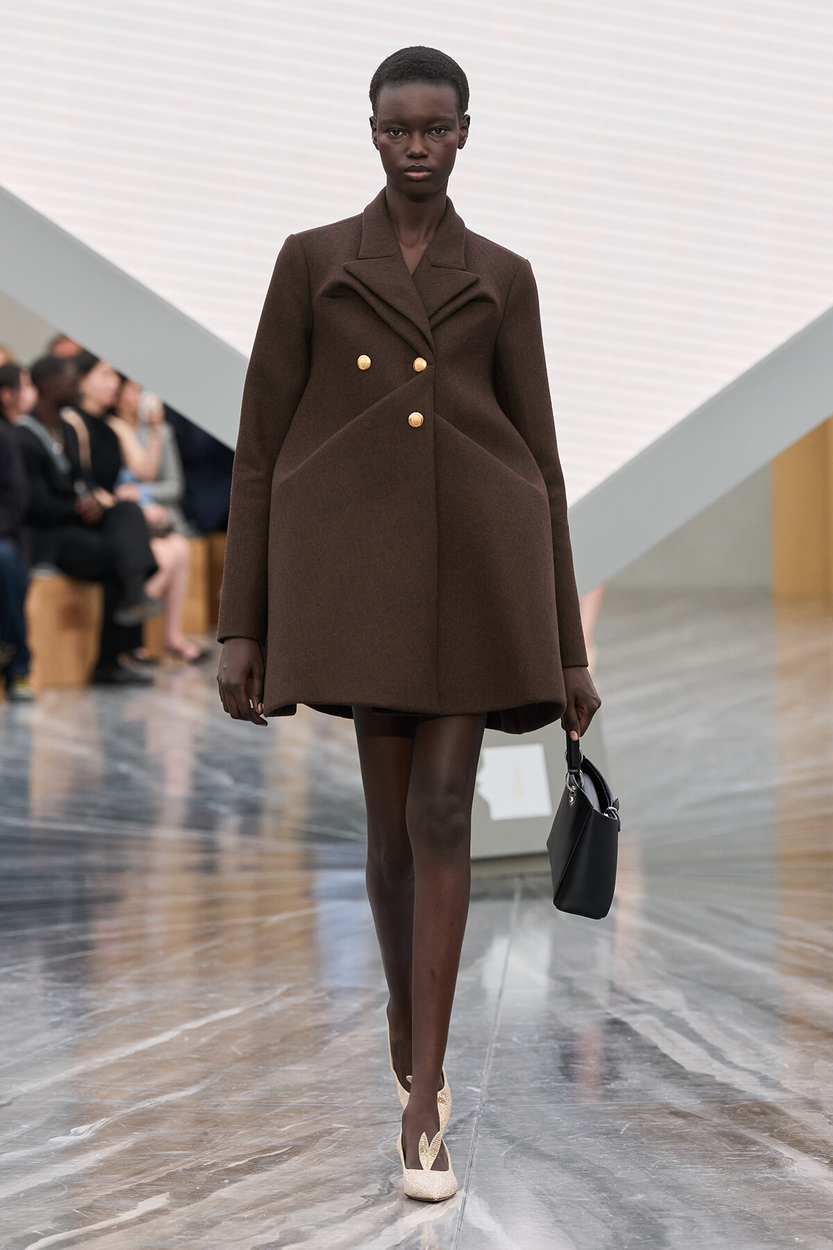 Model walks the runway in a brown structured double-breasted coat with gold buttons, carrying a small black handbag and wearing beige heels.