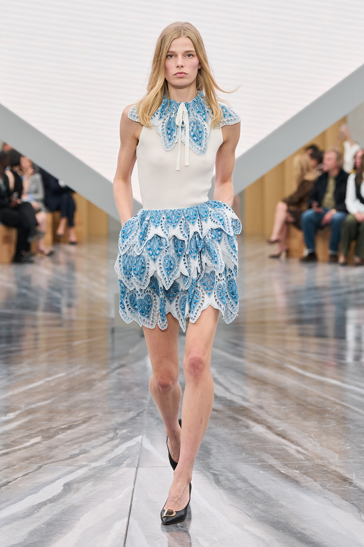 Model walking a runway in a white sleeveless top with a blue lace collar and a tiered blue lace skirt.