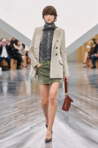Model walking a fashion show in a light beige blazer over a gray ruffled blouse with a bow, olive mini skirt, and burgundy heels, carrying a brown textured handbag.