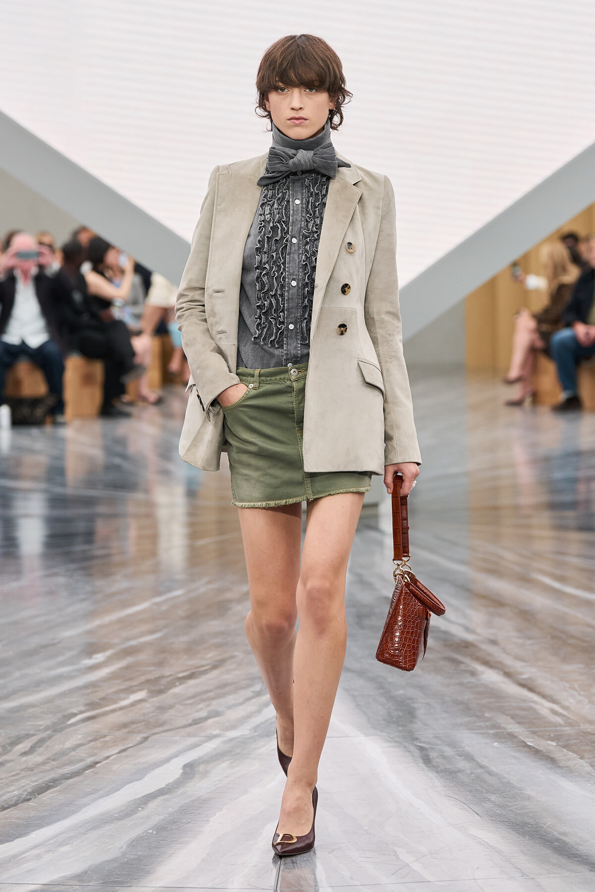 Model walking a fashion show in a light beige blazer over a gray ruffled blouse with a bow, olive mini skirt, and burgundy heels, carrying a brown textured handbag.