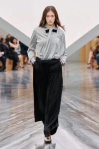 Model on a fashion runway wearing a gray blouse with a black bow and black wide-leg pants, hands in pockets.