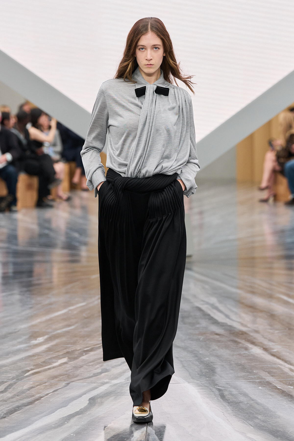 Model on a fashion runway wearing a gray blouse with a black bow and black wide-leg pants, hands in pockets.