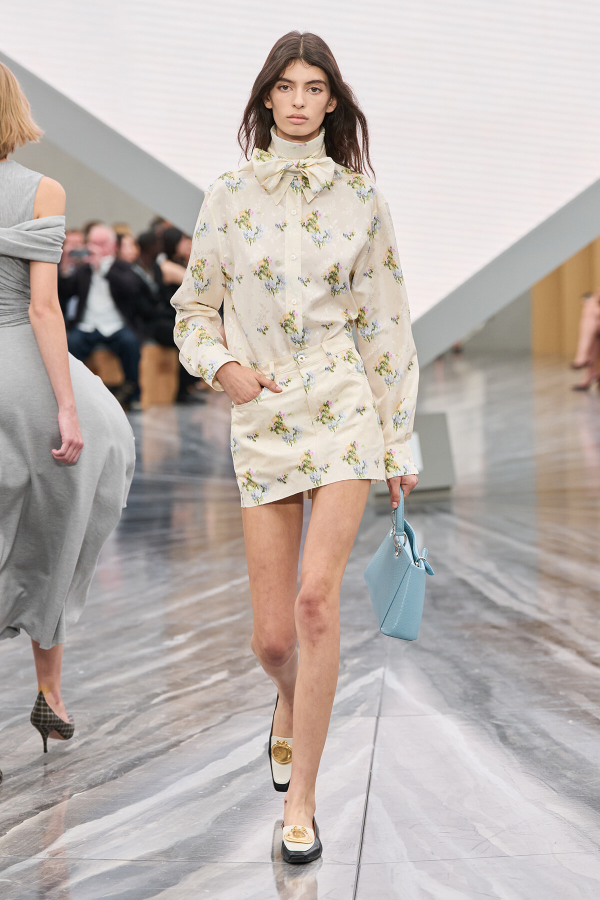 Model walking a runway in a cream floral shirt dress with a large bow at the neck, carrying a light blue handbag.