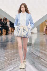 Model walking a runway in a pastel blue button-down shirt with a cream belt and a fluffy blue-and-white mini skirt.