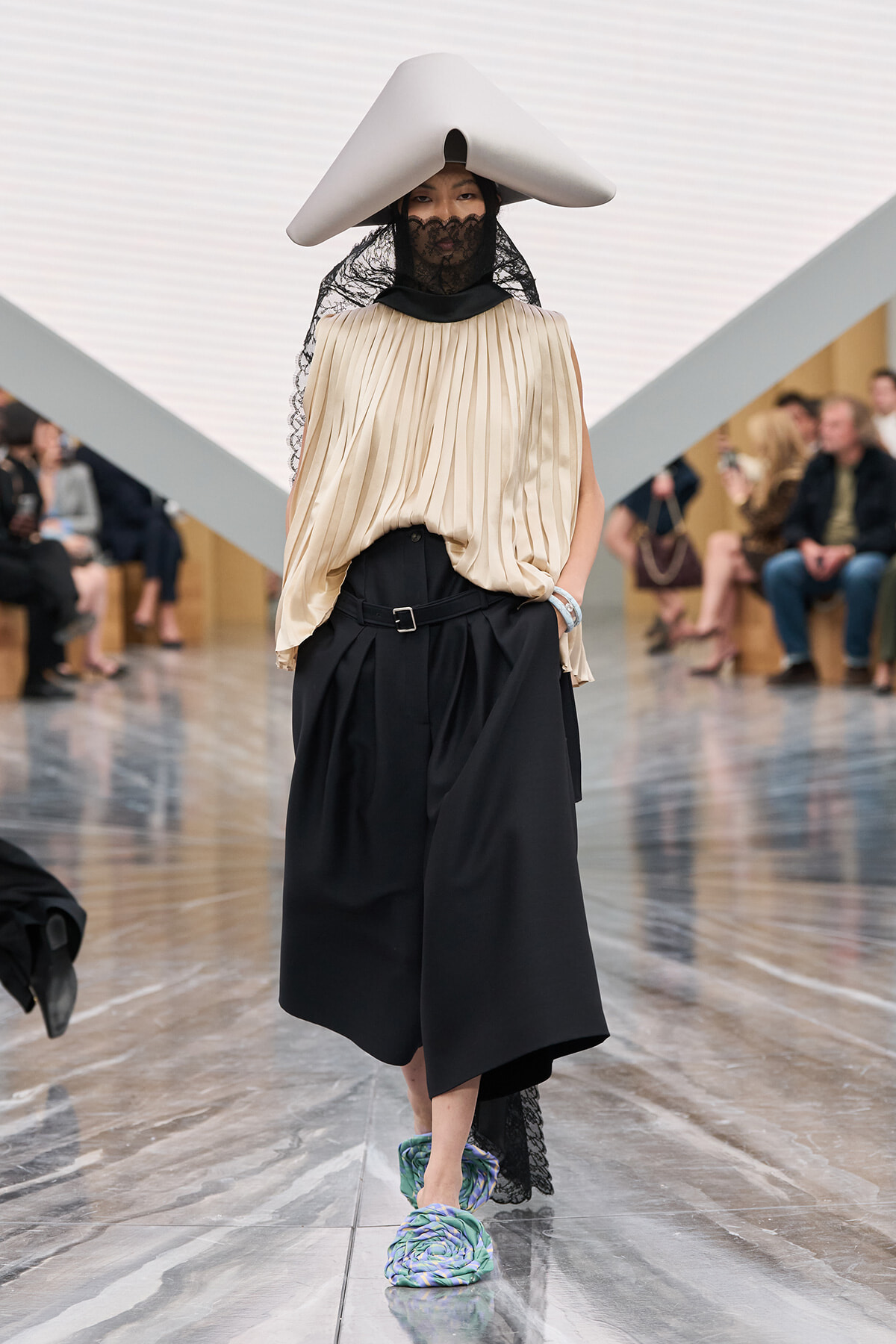Model walking a fashion runway in a beige pleated top, black wide-leg skirt with belt, a sculptural gray hat and lace veil, plus teal patterned shoes.