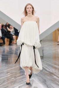 Model walks down a runway in a loose, white gown with gathered hem and oversized floral-embellished sleeves tied with black ribbons.
