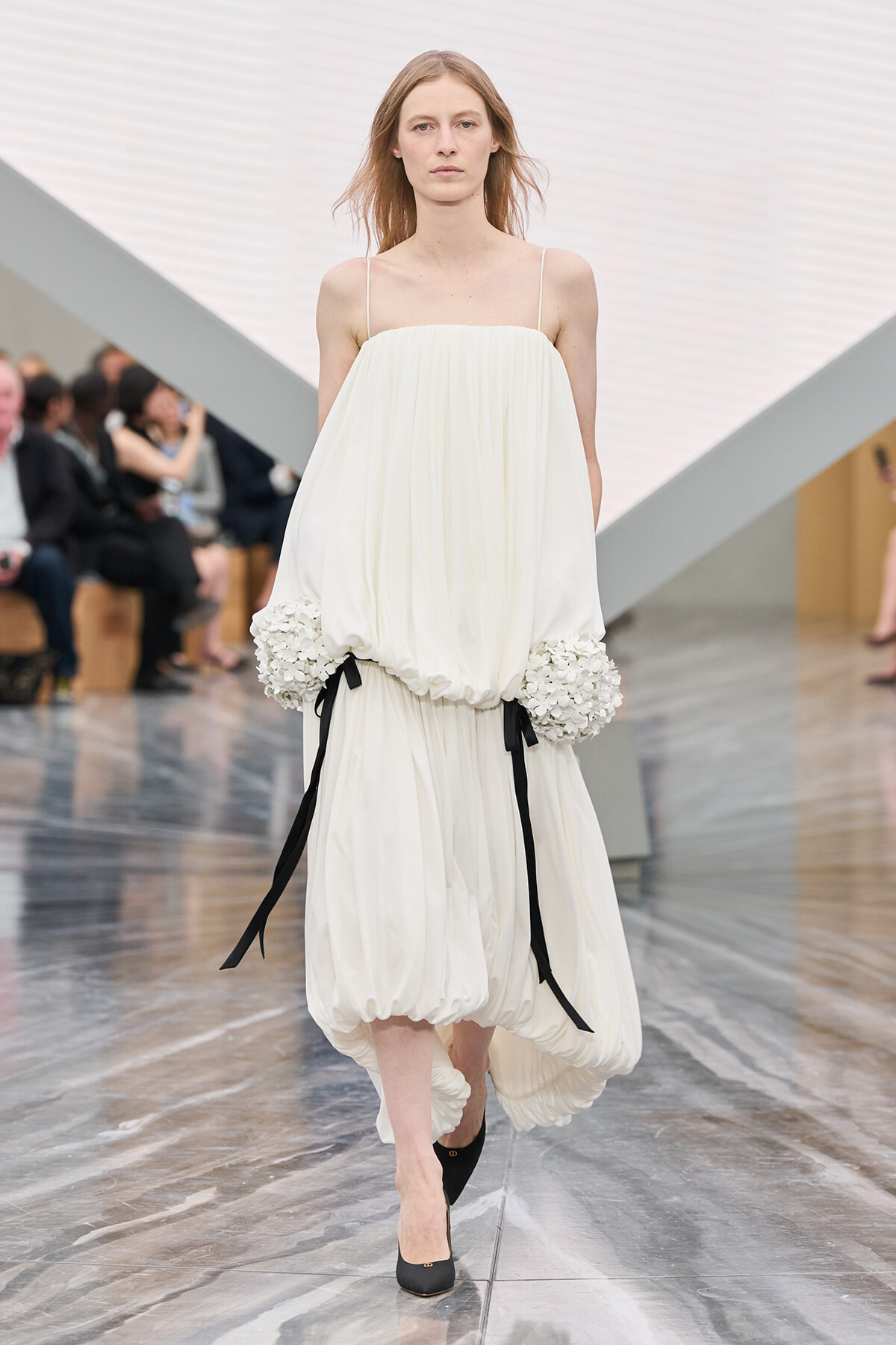 Model walks down a runway in a loose, white gown with gathered hem and oversized floral-embellished sleeves tied with black ribbons.