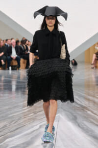 Female model walks a fashion runway in an all-black outfit with a sculptural wide-brim hat and a voluminous scalloped skirt, carrying a beige textured clutch.