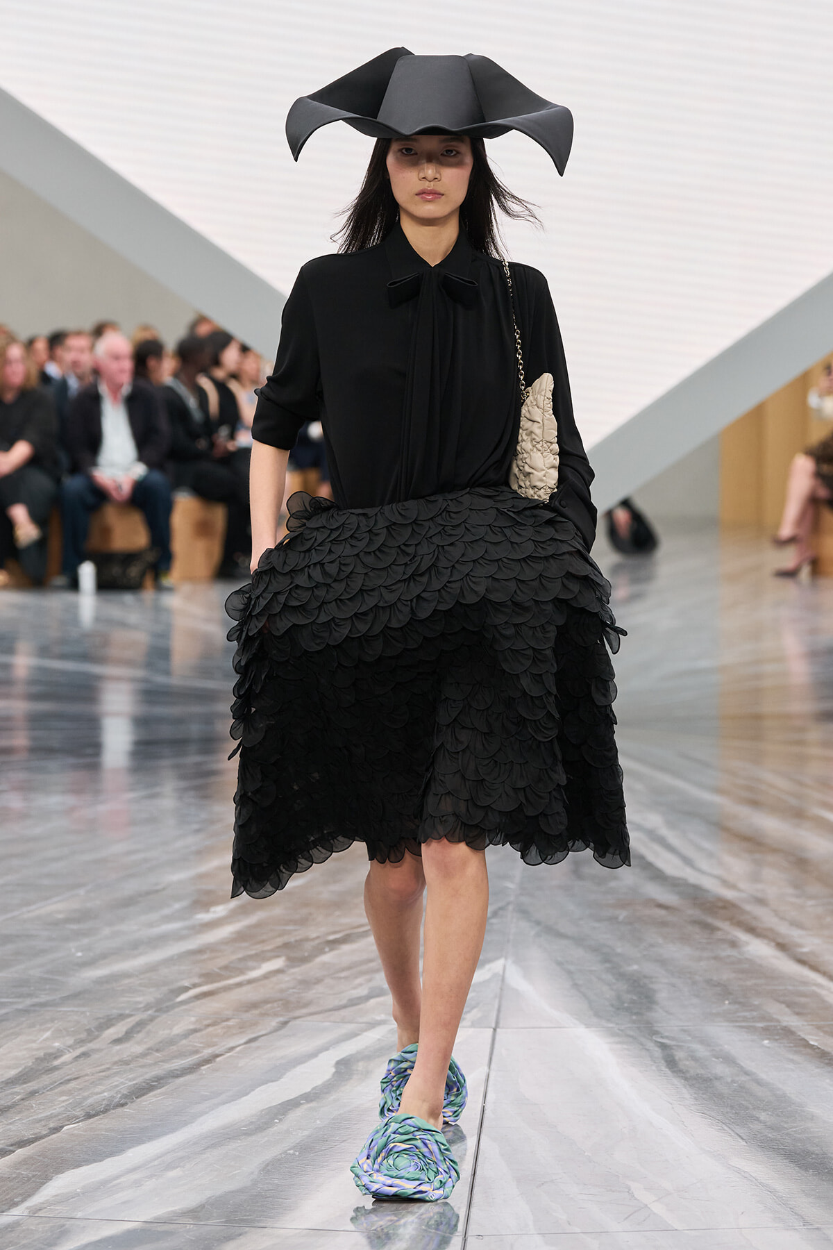 Female model walks a fashion runway in an all-black outfit with a sculptural wide-brim hat and a voluminous scalloped skirt, carrying a beige textured clutch.