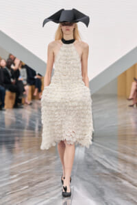Model walking a runway in a cream scalloped fringe dress with a black choker and an oversized black sculptural hat while spectators watch.