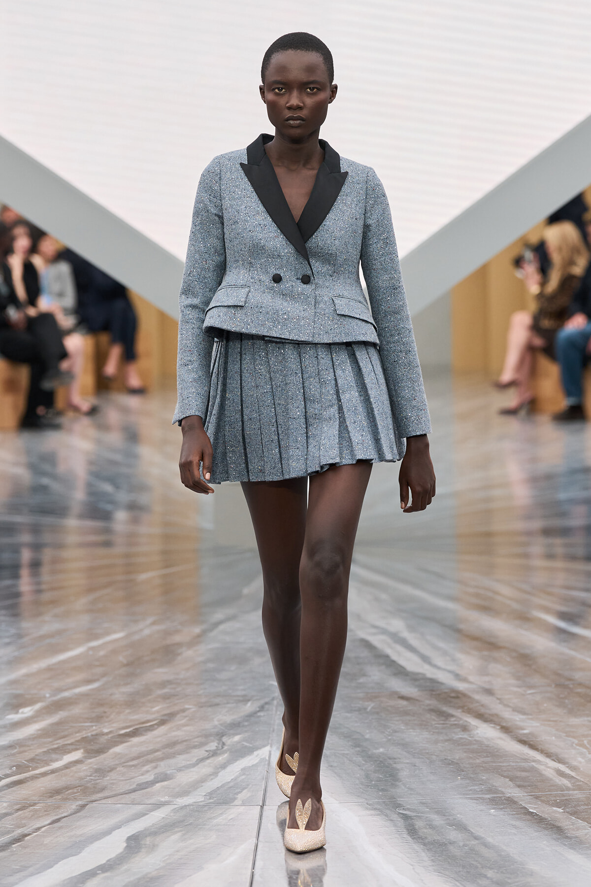 Female model on a runway wearing a shimmering blue-gray tailored blazer with black lapels and a matching pleated mini-skirt, gold glitter pumps visible.