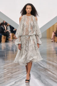 Model walking a runway in a pale silver lace gown with sheer overlay and voluminous sleeves, paired with black heels.