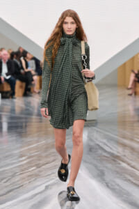 Model walking a fashion show in a green checkered dress with a bow at the neck, carrying a beige shoulder bag and wearing black loafers.