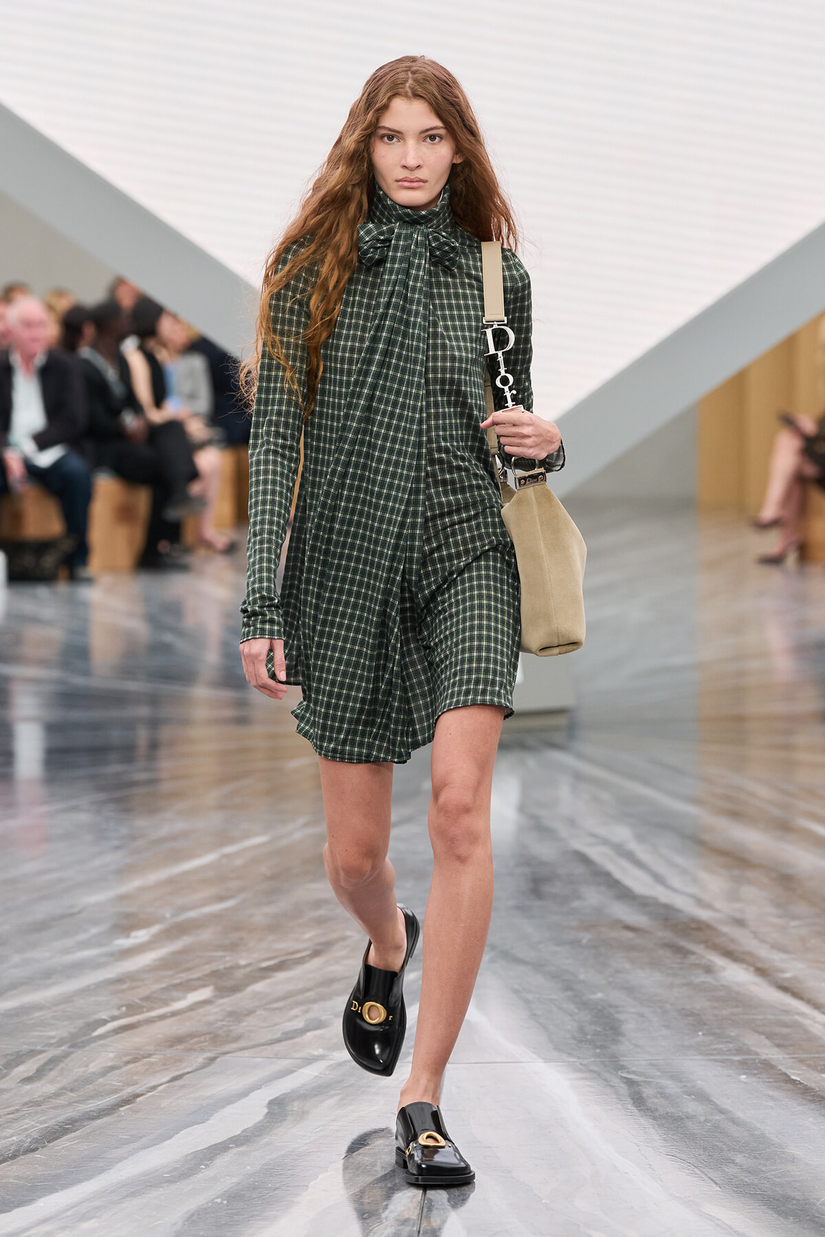 Model walking a fashion show in a green checkered dress with a bow at the neck, carrying a beige shoulder bag and wearing black loafers.