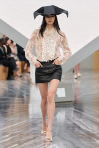 Model walking a runway in a sheer floral blouse with a ruffled front, black mini skirt, beige heeled sandals, and a sculptural black hat.