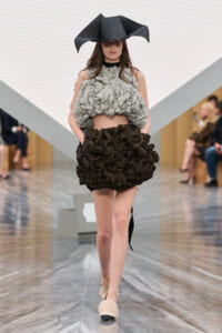 Model walks the runway in a voluminous gray ruffled crop top and a black ruffled mini skirt, wearing a large black sculptural hat.