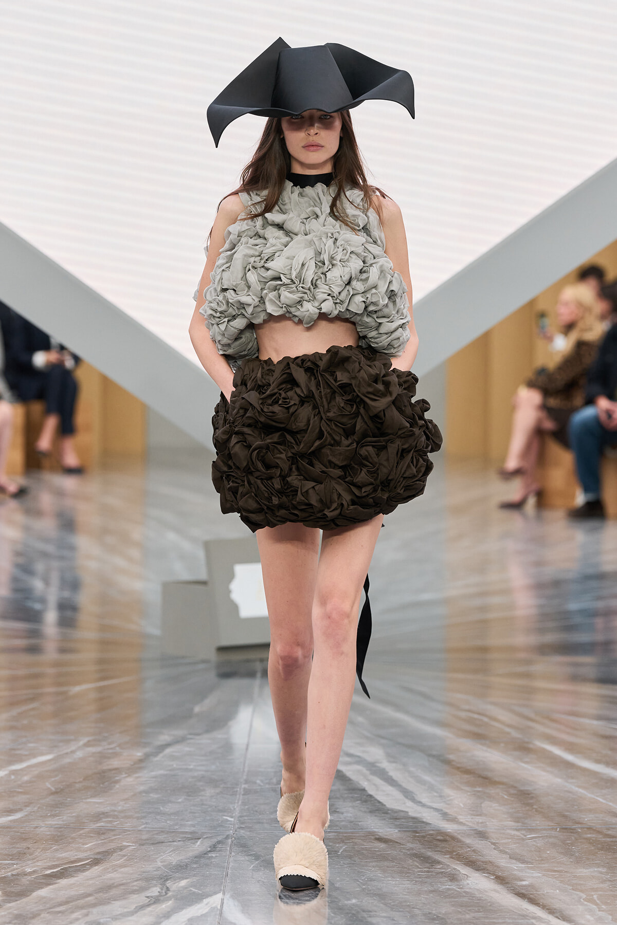 Model walks the runway in a voluminous gray ruffled crop top and a black ruffled mini skirt, wearing a large black sculptural hat.