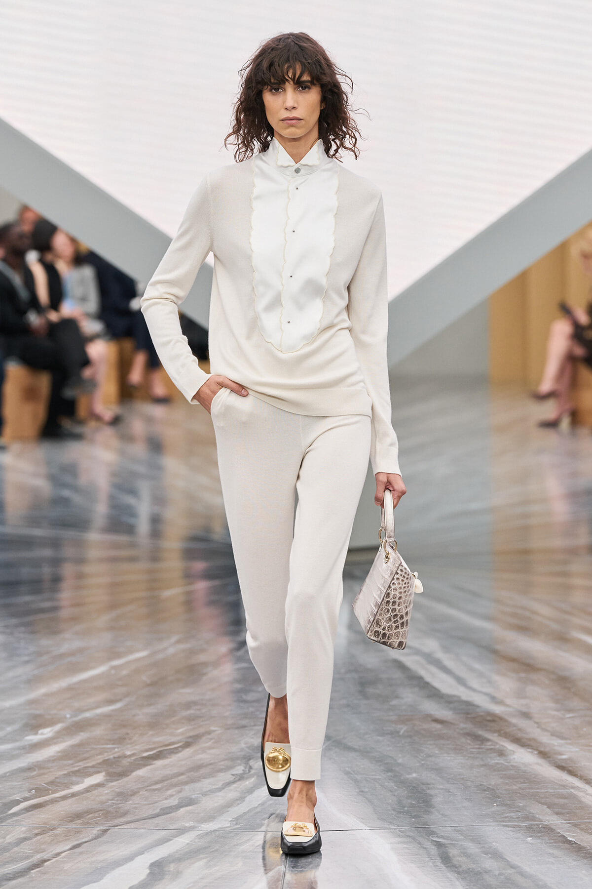 Model walks a cream suit with a ruffled front blouse on a glossy runway, carrying a pale crocodile-pattern bag.