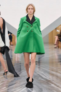 Model walking runway in bright green double-breasted coat with black lapels and gold buttons, black ruched slip-on shoes.