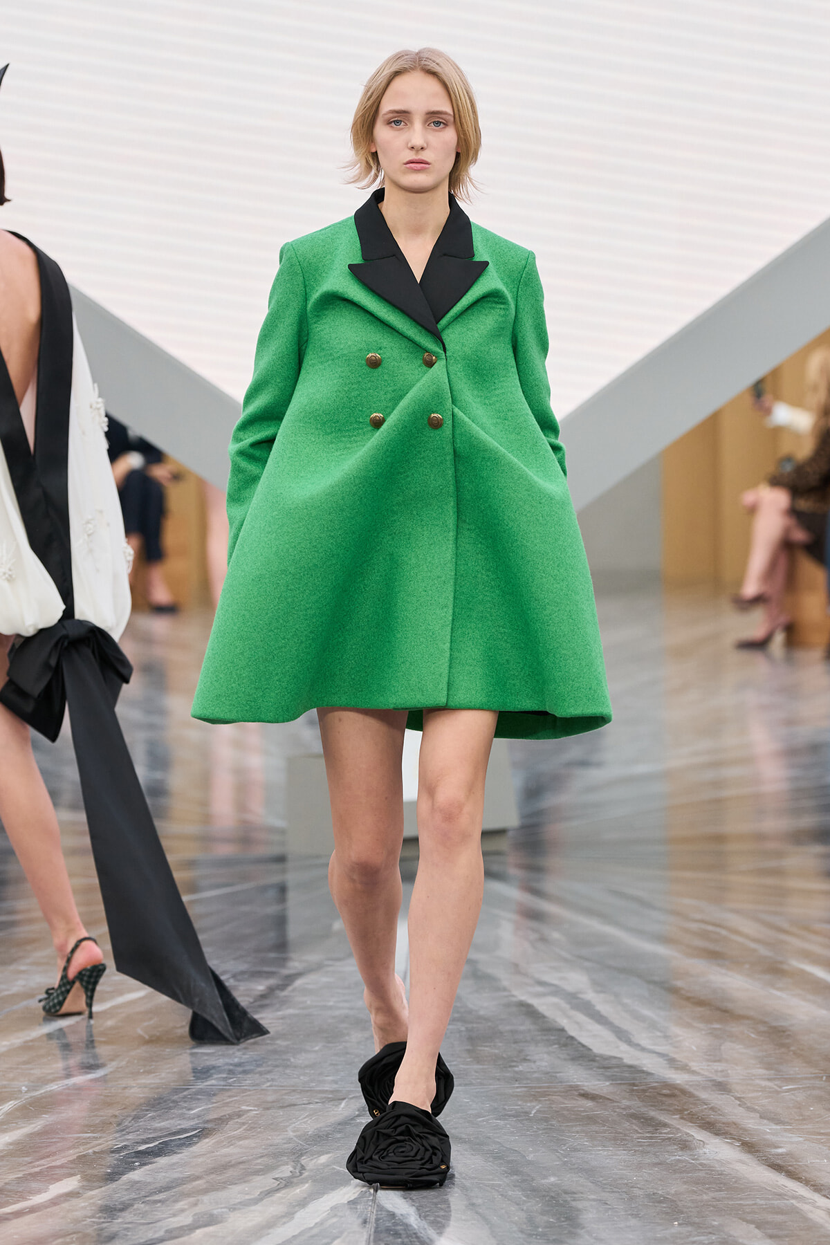 Model walking runway in bright green double-breasted coat with black lapels and gold buttons, black ruched slip-on shoes.