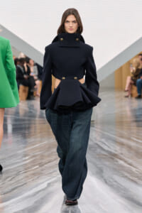 Female model walks the runway in a dark navy sculptural coat with a high collar and peplum, worn over loose denim jeans.