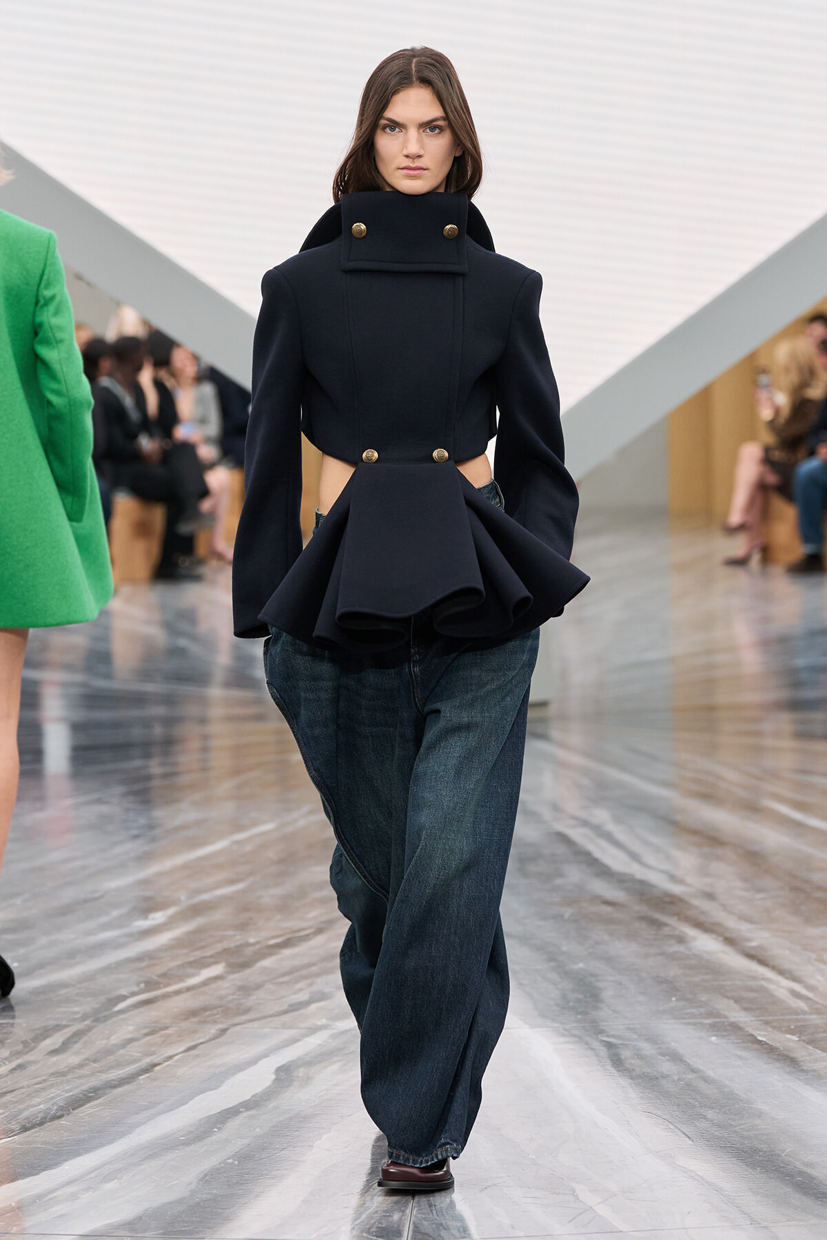 Female model walks the runway in a dark navy sculptural coat with a high collar and peplum, worn over loose denim jeans.